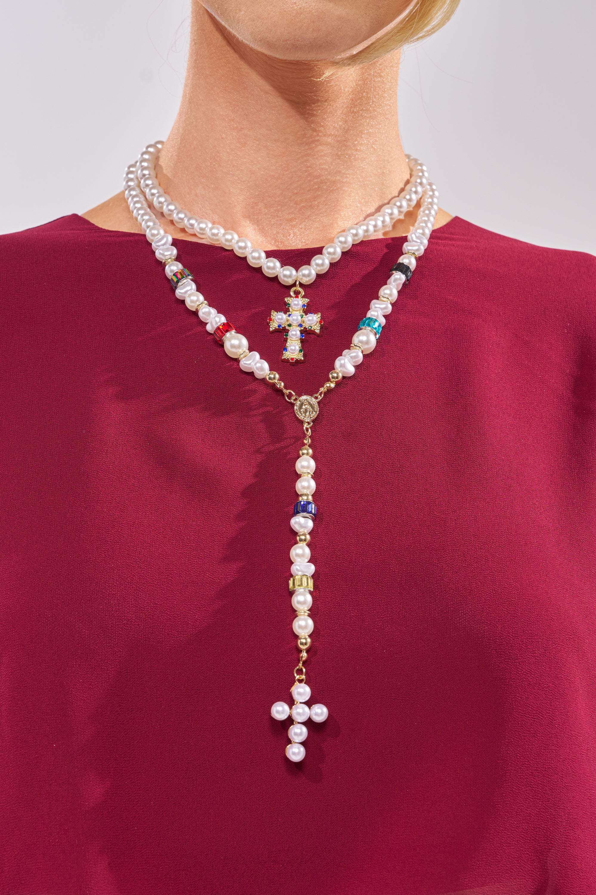 Wearing a burgundy top, the person showcases the ON BISCAYNE NECKLACE—a pearl necklace set with two faux pearl strands. Each cross pendant features pearls and colorful beads, elegantly resting on the upper chest and neck. 1