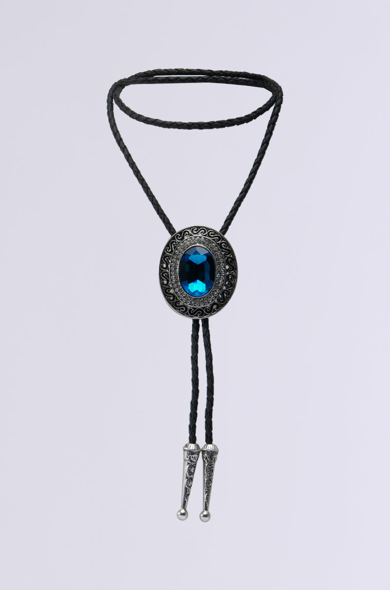 The QUEEN BOLO TIE NECKLACE is a black braided necklace with silver tips. It boasts an ornate oval clasp made of silver, featuring intricate detailing. At the center is a large, vertically oriented dark blue gemstone, surrounded by subtle rhinestone accents against a plain light gray background.
