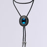 The QUEEN BOLO TIE NECKLACE is a black braided necklace with silver tips. It boasts an ornate oval clasp made of silver, featuring intricate detailing. At the center is a large, vertically oriented dark blue gemstone, surrounded by subtle rhinestone accents against a plain light gray background.