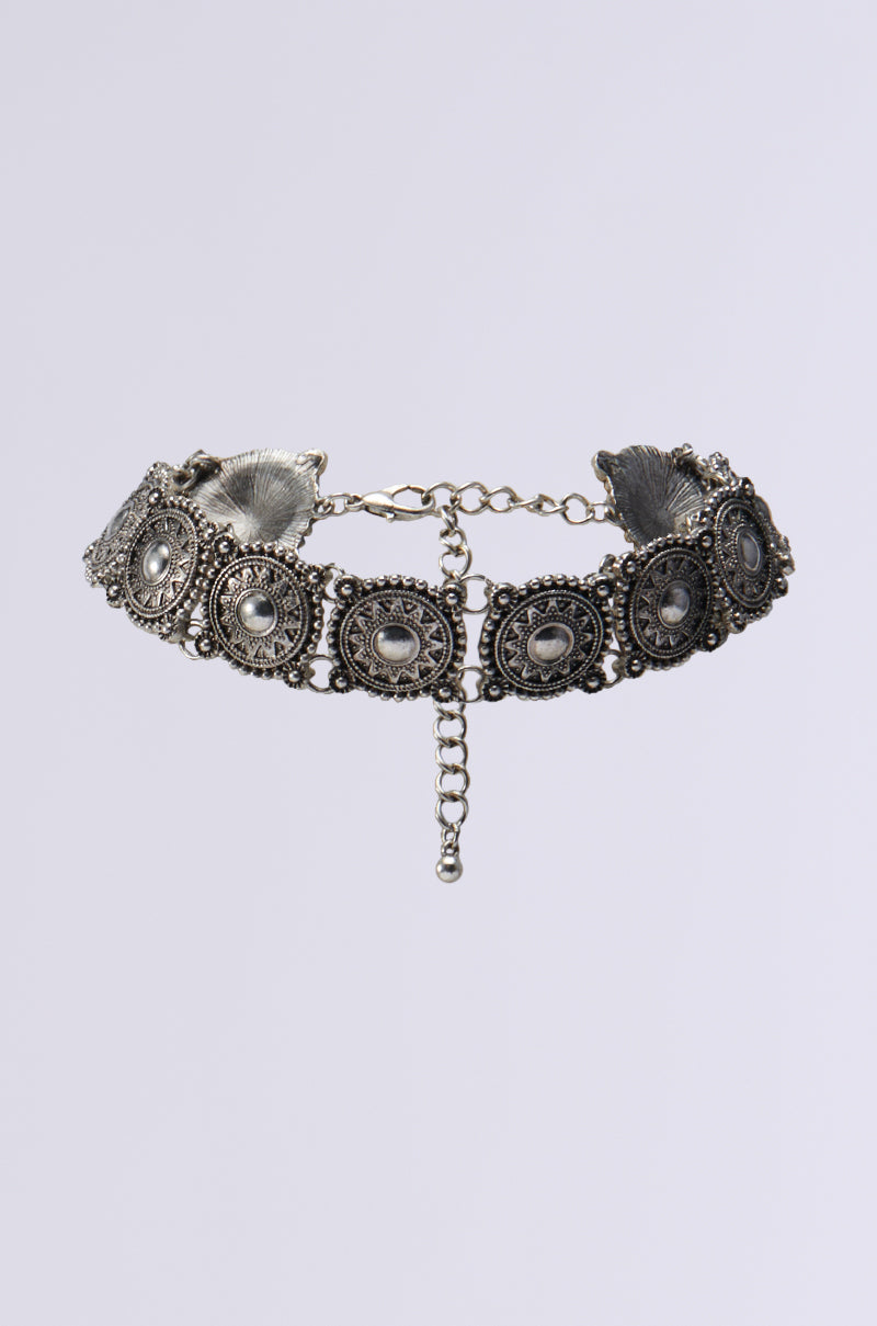 The WESTERN CHIC CHOKER is a vintage-style silver necklace adorned with intricate circular filigree designs, teardrop-shaped embellishments, and Western-inspired medallions. Its adjustable lobster clasp closure guarantees a perfect fit and adds an elegant, classic touch when worn against a neutral background.