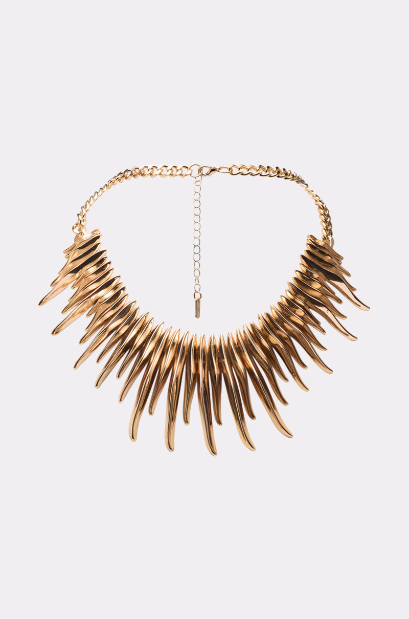 The UNPREDICTABLE NECKLACE features gold-tone elongated spikes in a fan design, attached to a gold chain with an adjustable clasp, showcased on a plain light background. 0