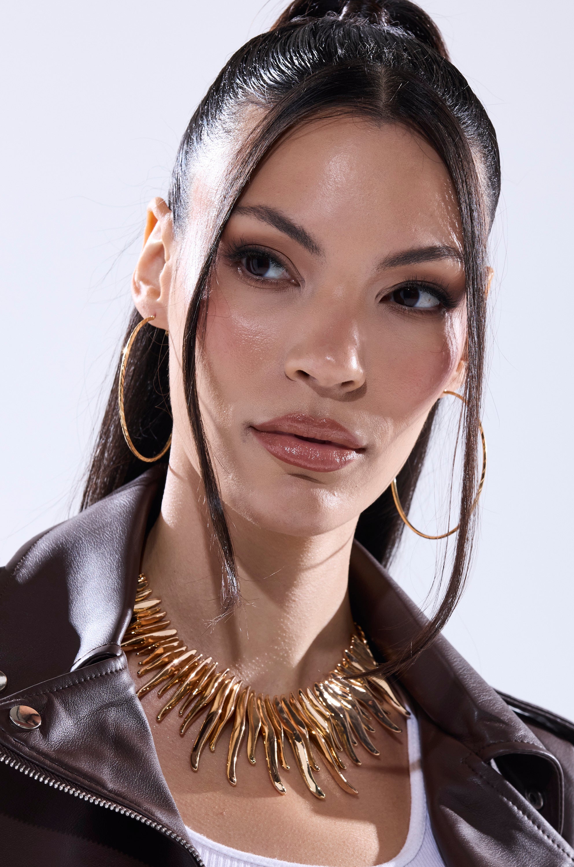 A woman with long dark hair in a high ponytail wears gold hoop earrings, a white top, a brown leather jacket, and the UNPREDICTABLE NECKLACE. She gazes slightly to the side against a light background. 3
