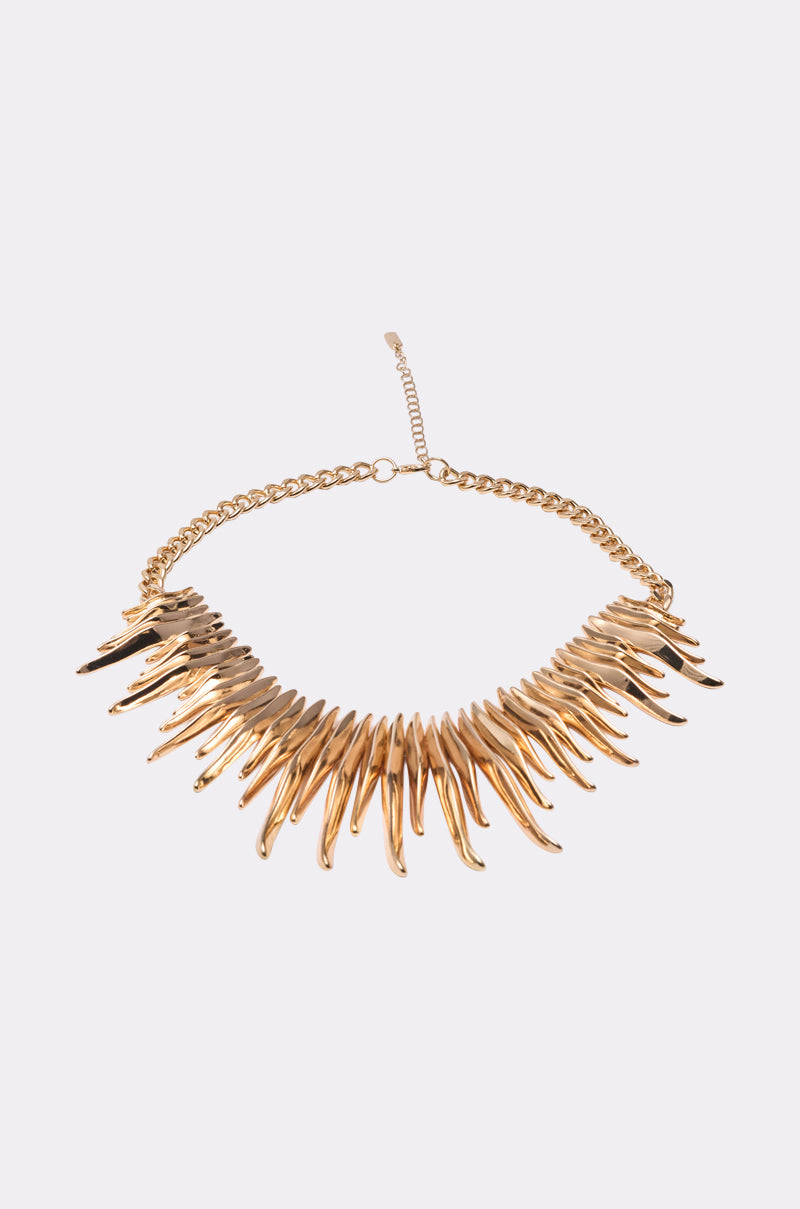 The UNPREDICTABLE NECKLACE is a gold-tone statement piece with pointed, flame-like spikes radiating from a gold chain and adjustable clasp, shown against a light background. 2