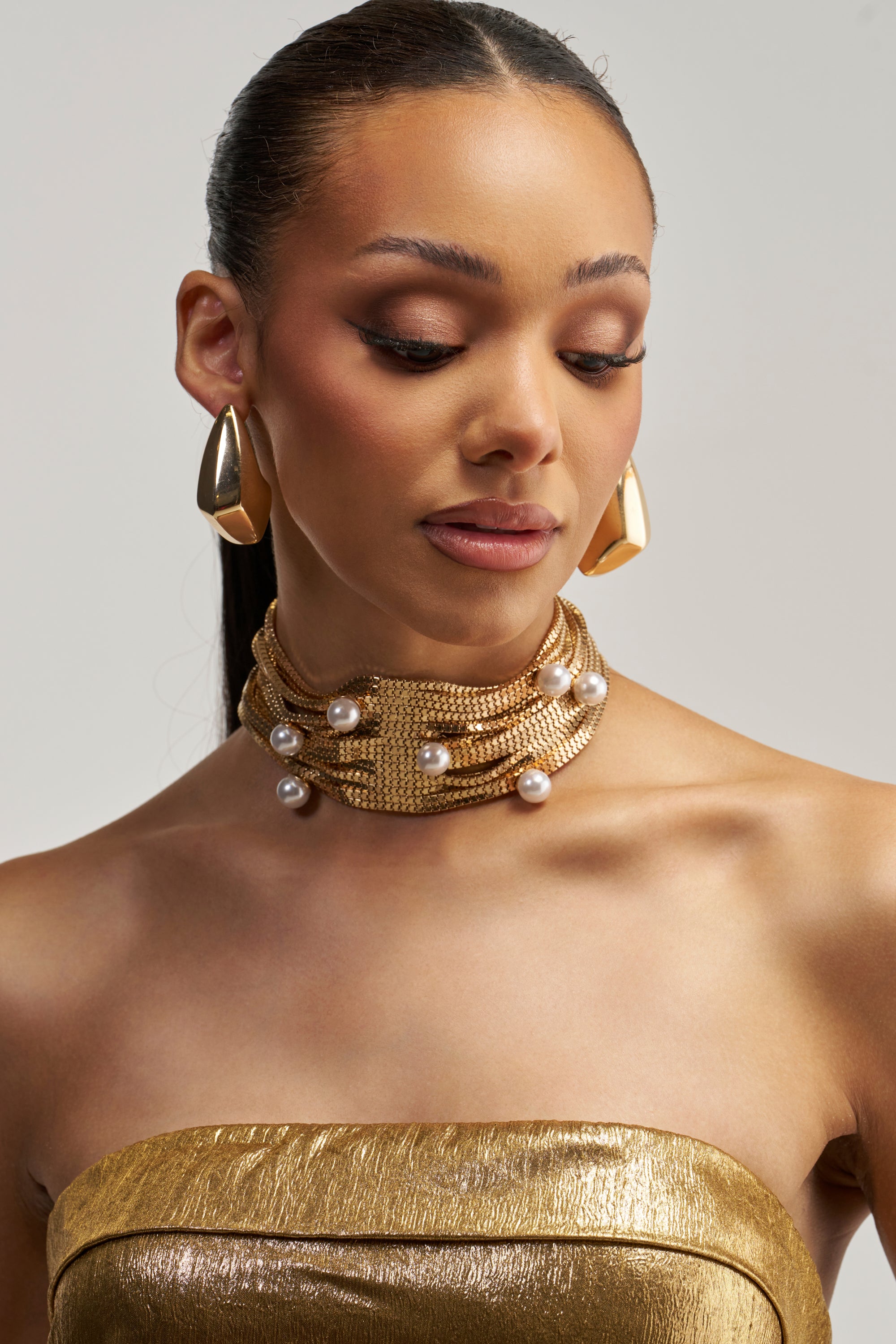 A woman with a sleek ponytail wears gold earrings and the PEARL PERFECTION NECKLACE, a statement collar adorned with pearls, paired with a gold strapless top. She gazes downward serenely against a plain background.