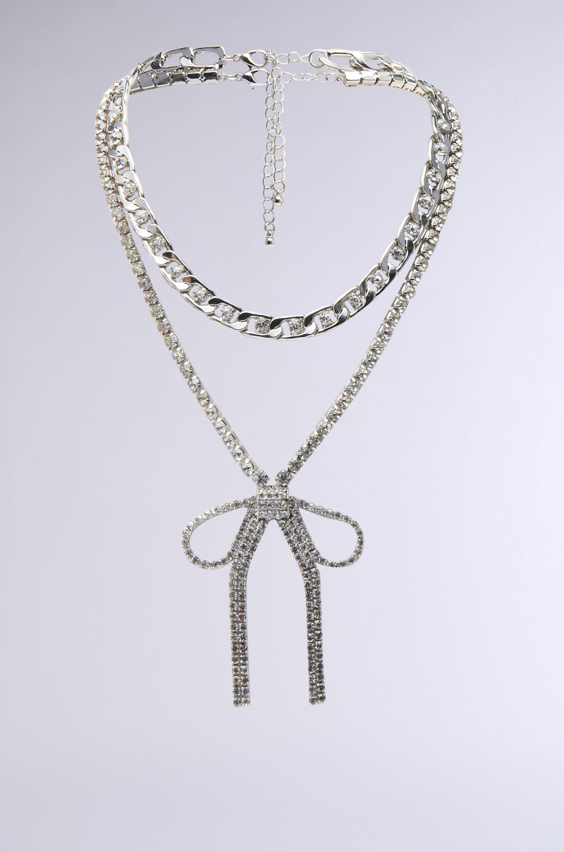 The WITH A BOW SILVER NECKLACE SET features a chunky chain, a slim chain with a sparkling bow pendant, and an adjustable extender clasp, shown on a light gray background. 0