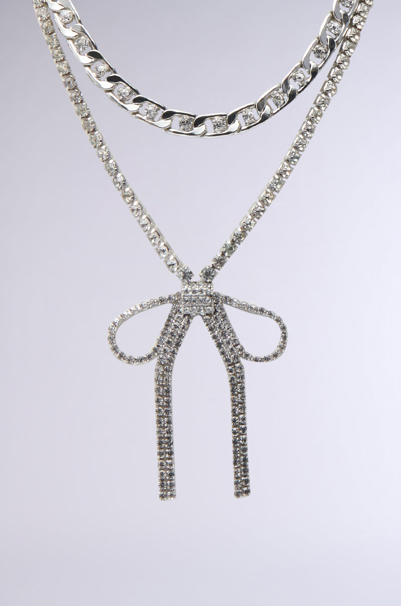 The WITH A BOW SILVER NECKLACE SET features two silver necklaces: a chunky chain and a slim necklace with a rhinestone bow pendant, displayed against a plain light gray background.