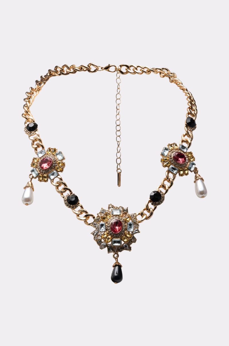 The DOLCE VITA NECKLACE is a gold chain statement piece with three jeweled floral pendants adorned with pink and clear stones, black beads, and pearl-like drops, elegantly displayed against a light background. 0
