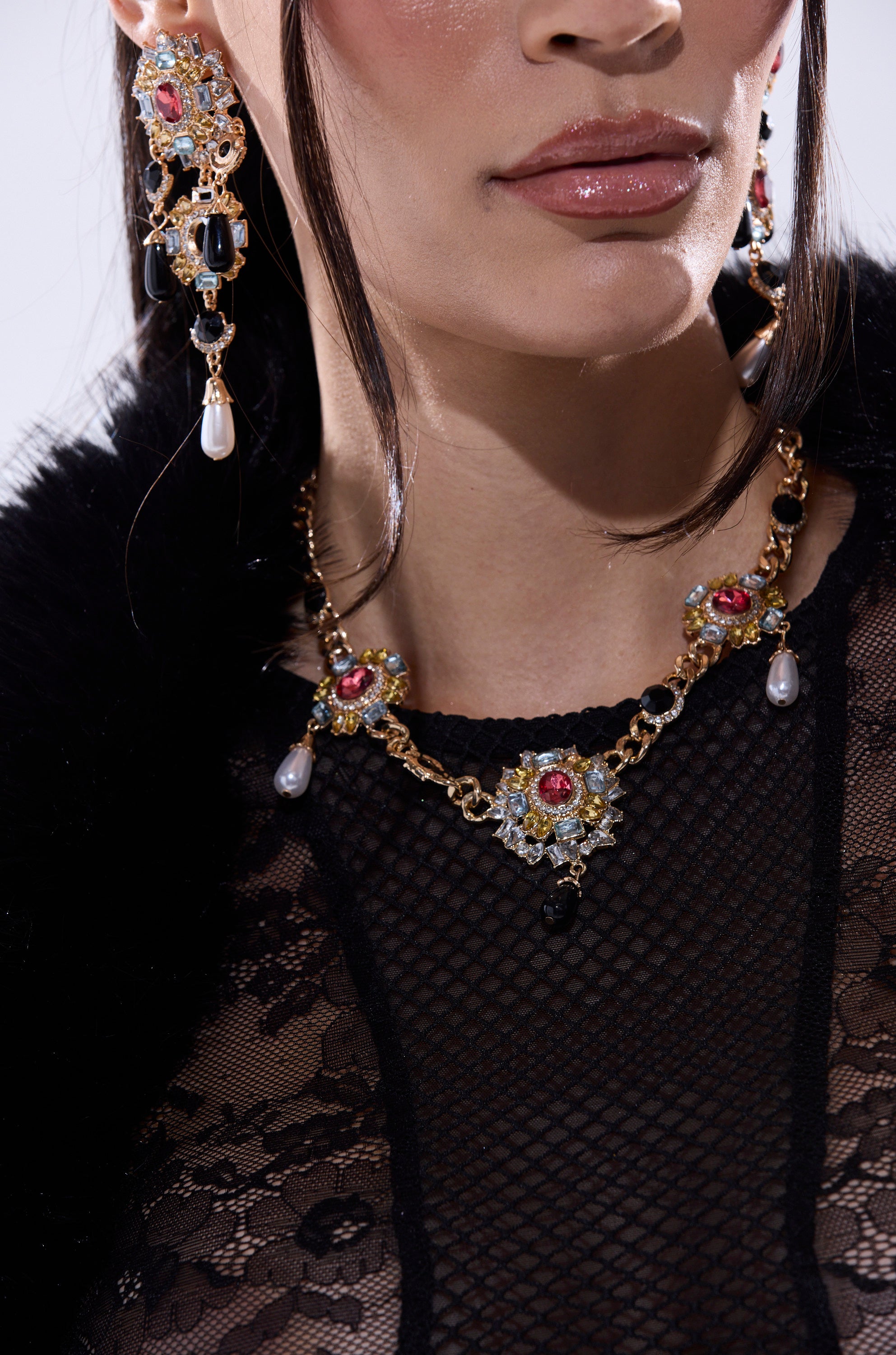 A woman wears the DOLCE VITA NECKLACE—ornate gold jewelry with pearls and gemstone clusters—and large earrings over a black lace mesh top. Only the lower half of her face and upper chest are visible. 1
