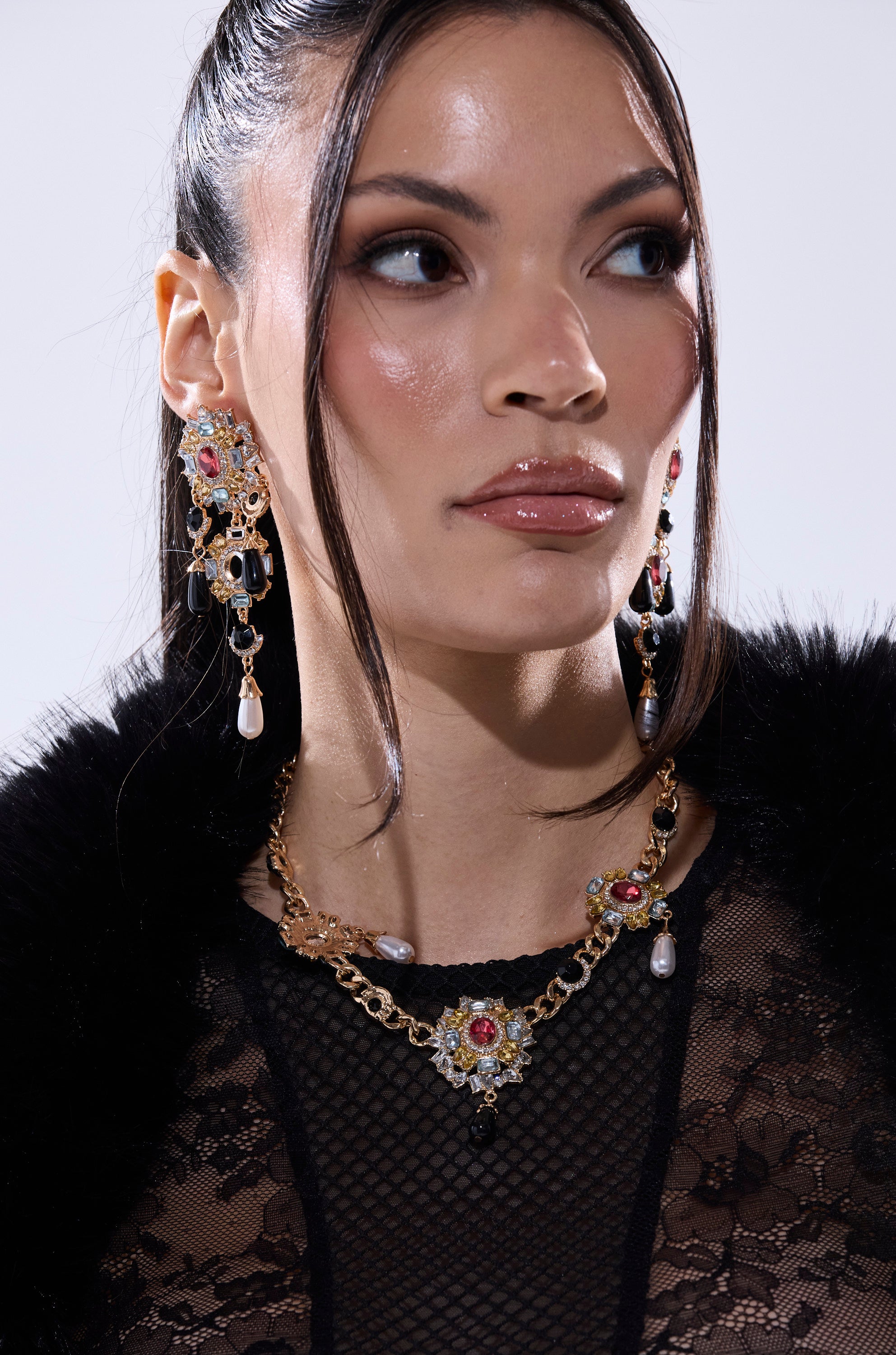 A woman with long dark hair in a sleek ponytail wears dramatic earrings, the DOLCE VITA NECKLACE, and a black lace top with fur trim. Her makeup is bold, featuring glossy lips and smoky eyes. 2