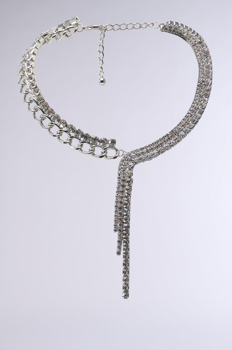 Introducing the WATERFALL BLING NECKLACE, a dazzling silver creation featuring a stylish cuban chain link adorned with rows of shimmering rhinestones. An asymmetrical tasseled rhinestone fringe gracefully drapes from one side, elegantly showcased on a simple gray backdrop.