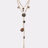 The HERE FOR THE VIBES NECKLACE features layered gold chains, textured metallic coin pendants, and a Y-shaped drop finished with a leaf charm, showcased on a simple light background.