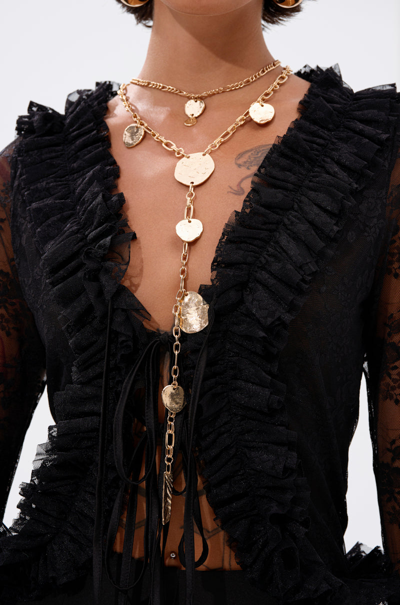 A person wearing a sheer black ruffled top with a deep neckline, accessorized with the HERE FOR THE VIBES NECKLACE and hoop earrings. The focus is on the statement necklace and ruffle details. 1