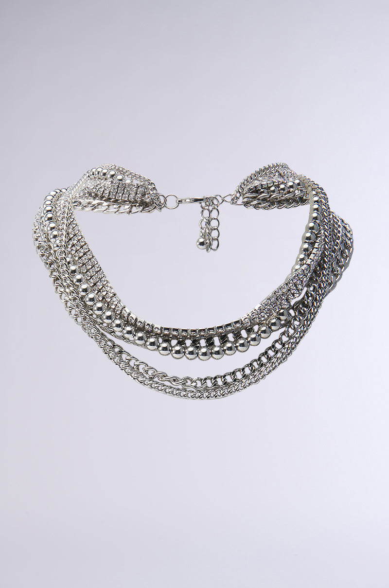 Introducing the ULTIMATE STACK LAYERED NECKLACE, a striking silver statement piece that boasts multiple chains and round metal beads, adorned with sparkling rhinestone embellishments and an adjustable clasp. It is beautifully showcased against a plain gray background. 0