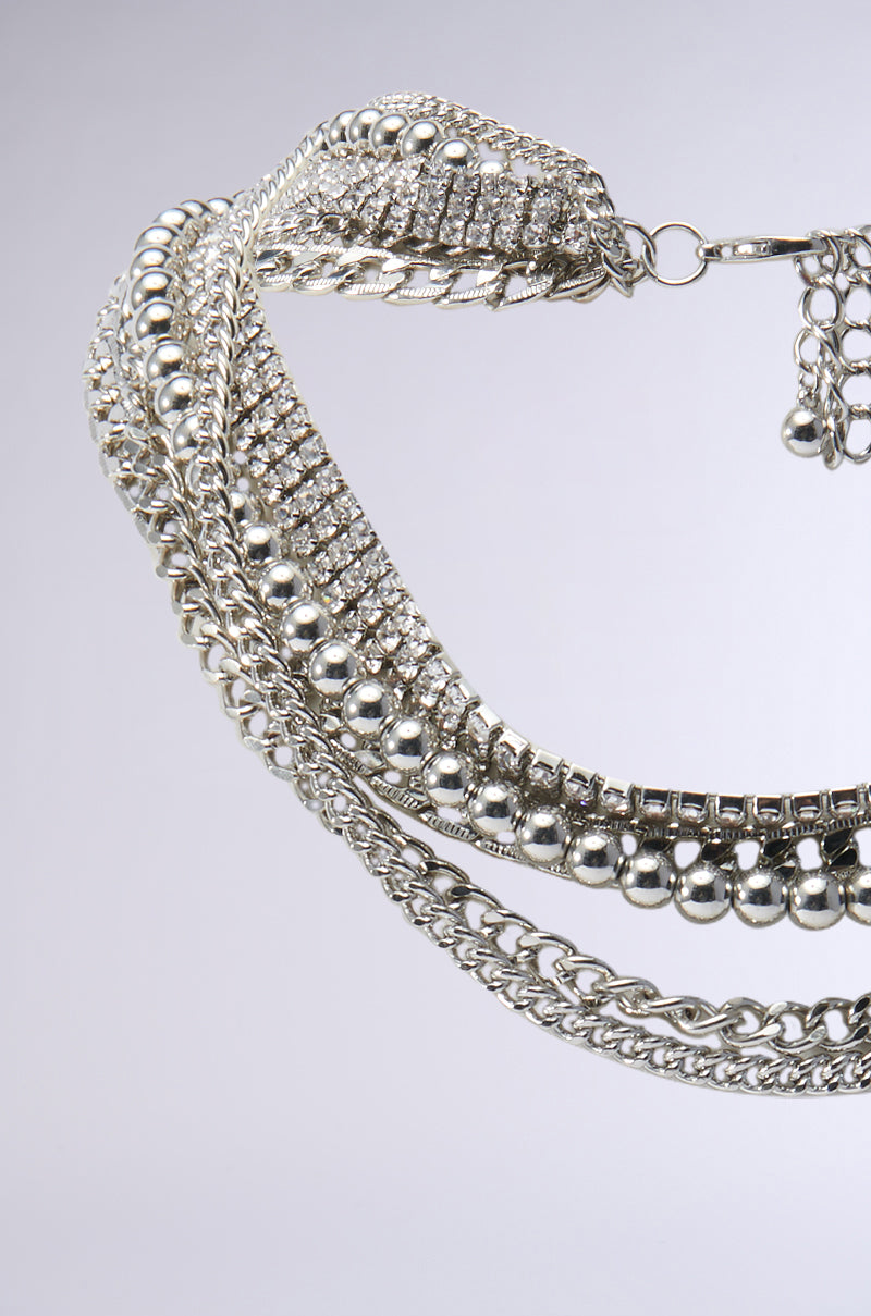 The ULTIMATE STACK LAYERED NECKLACE is featured in a close-up image, displaying its multi-strand silver design with an array of diverse chain styles. It includes round beads, textured links, and sparkling rhinestone embellishments that create an elegant overlapping pattern against a light gray background. 2