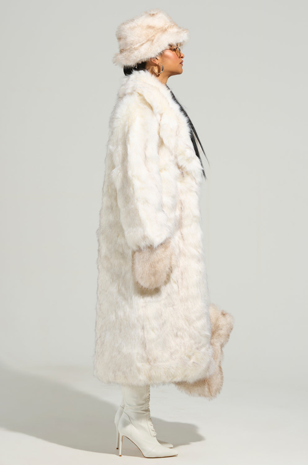 A person is striking a pose in profile, wrapped in the TOASTED MARSHMALLOW FUR COAT, along with a matching hat and white high-heeled boots. Their long dark hair cascades down past their shoulders, highlighted by chic sunglasses. They confidently tuck their hands into the coat's pockets against a plain gray backdrop.