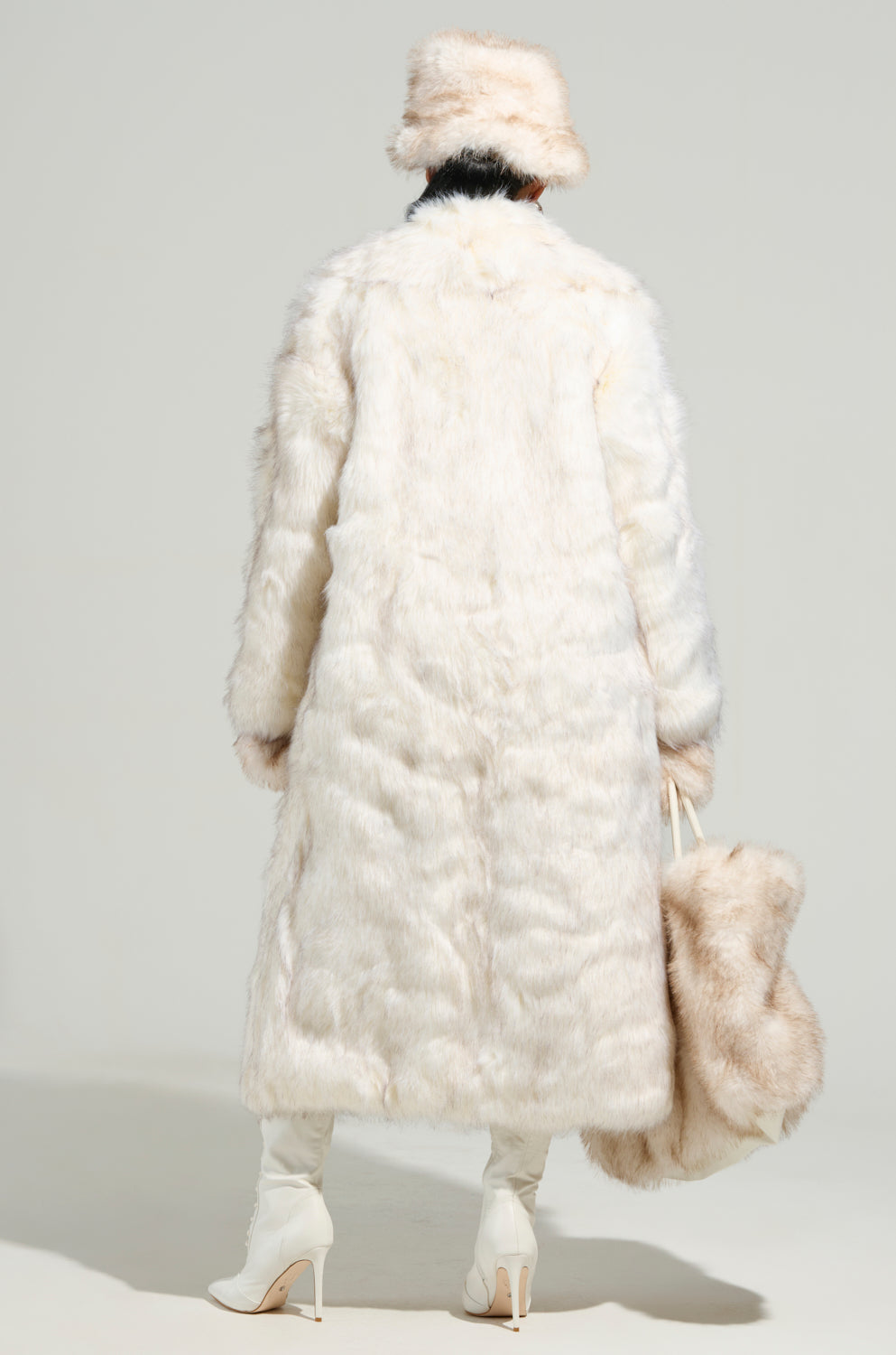 A person stands with their back to the camera, wrapped in an elegant TOASTED MARSHMALLOW FUR COAT and a coordinating hat. They hold a large, fluffy bag while wearing white high-heeled boots. The faux fur of the outfit forms an opulent maxi-length silhouette against the simple gray backdrop.
