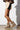 A person wearing a brown puffer jacket, creamy ruffled skirt, black sleeve on one arm, and the AZALEA WANG TOASTY BROWN BOOTIE with a faux suede upper stands against a white wall. The outfit combines textures and neutral tones for a stylish, cozy look.