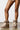 A person stands on a polished floor, wearing the AZALEA WANG TOASTY BROWN BOOTIE, featuring thick white laces and a faux suede upper. The well-lit background is plain, accentuating the textured flat sole of the boots and the person's legs.