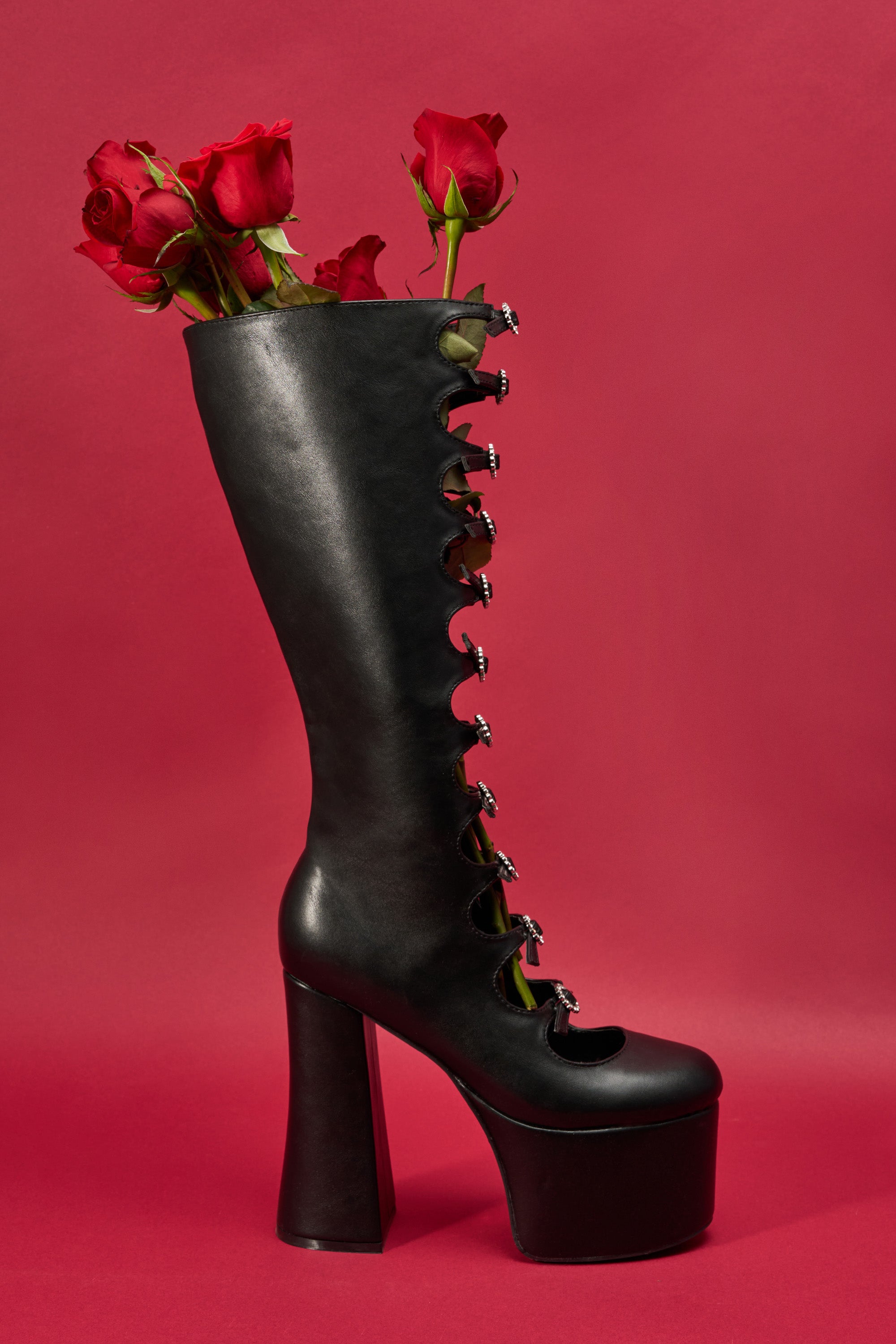 The AZALEA WANG TOMLIN BLACK BOOT, featuring decorative silver eyelets and a bold platform sole, stands upright against a red background with several red roses emerging from the top.