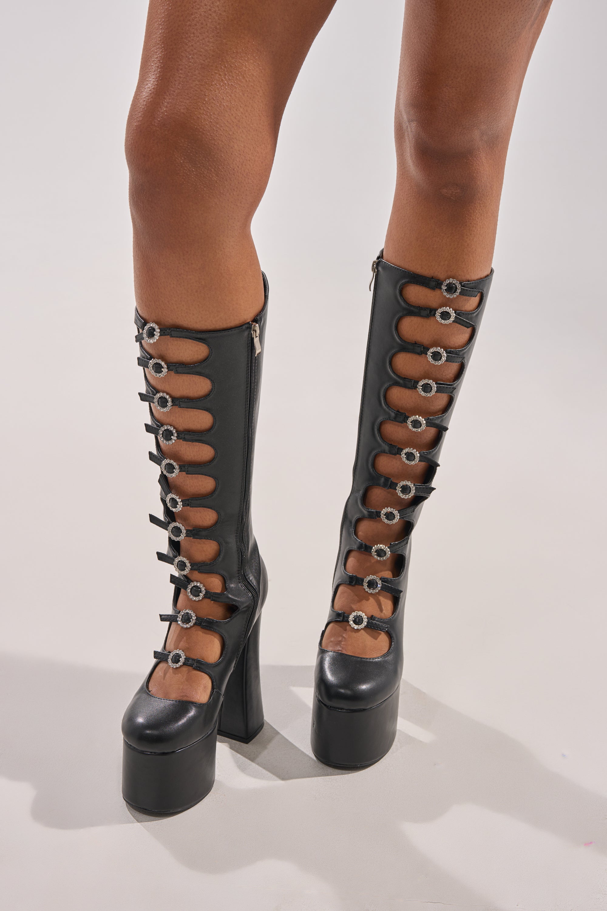 A person wears AZALEA WANG TOMLIN BLACK BOOT—knee-high, black boots with platform soles, multiple cut-outs, and silver buckle details—standing on a light background.