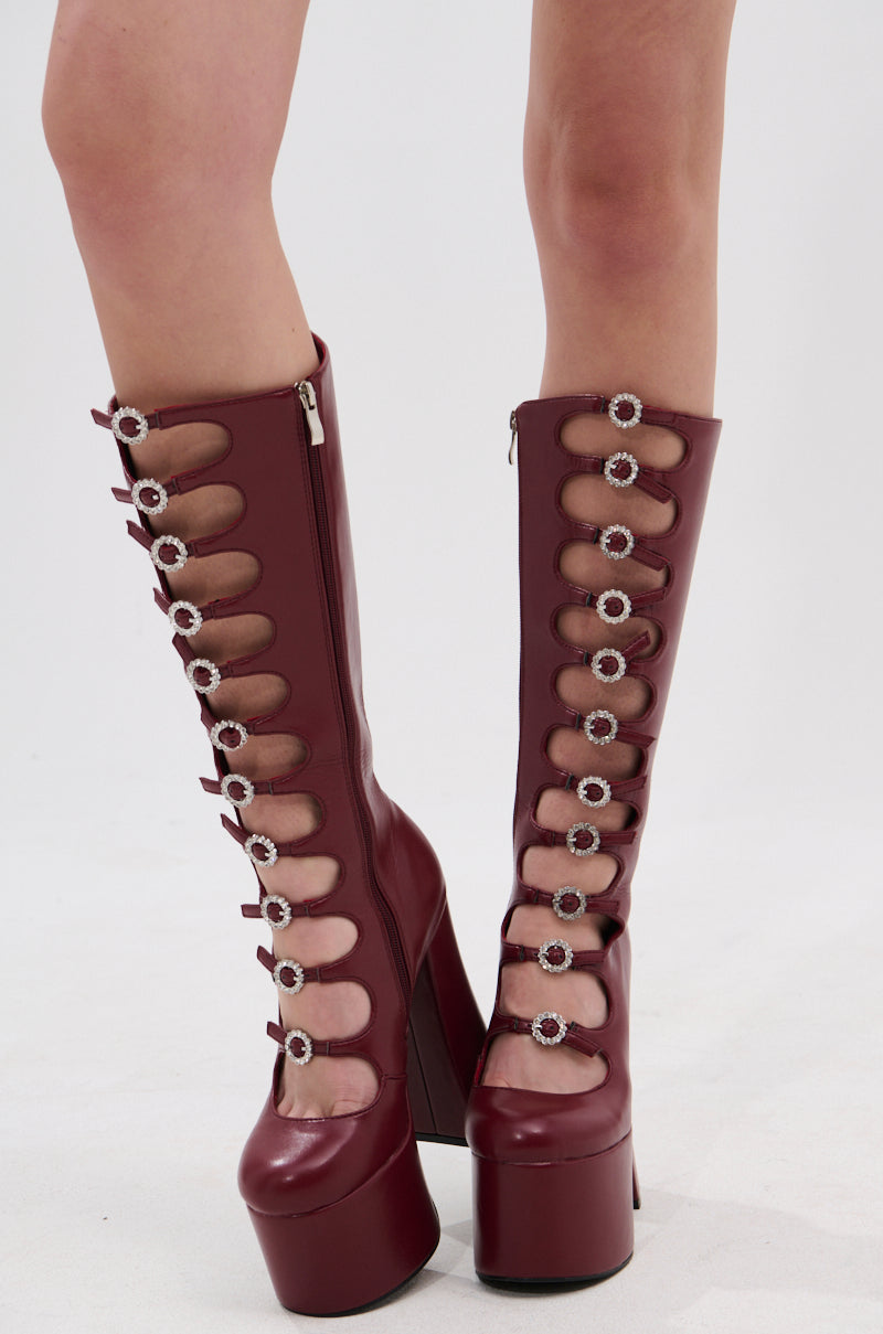Someone stands on a white background wearing the AZALEA WANG TOMLIN Burgundy Platform Boot—tall faux leather boots with multiple cutouts, round decorative buckles, and chunky heels.