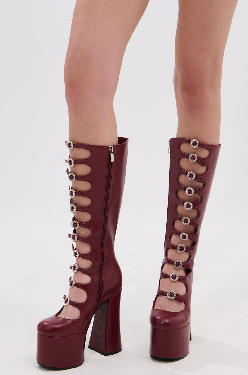 A person wearing the AZALEA WANG TOMLIN BURGUNDY PLATFORM BOOT—knee-high faux leather boots with multiple circular cutouts, jeweled buckles, and chunky heels—stands on a white background.
