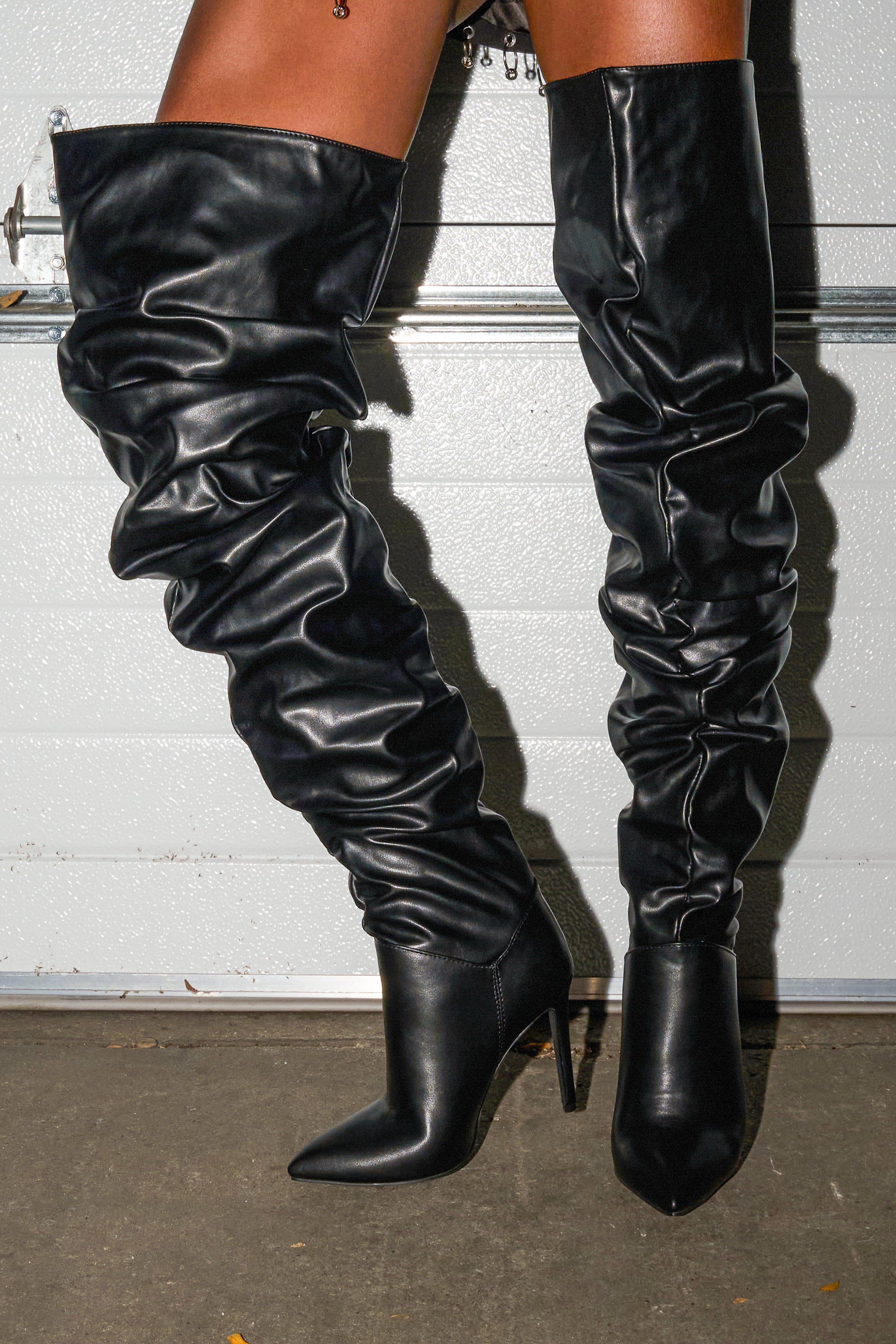A person wearing AZALEA WANG TORVIS Ruched Stiletto Boots in black stands on a concrete floor in front of a metallic garage door.