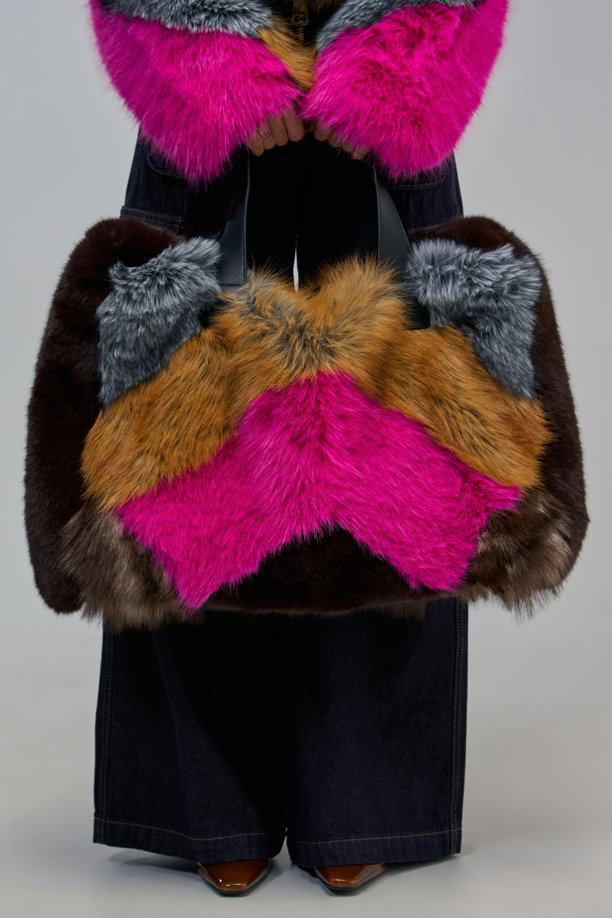 WERE ALL MAD HERE OVERSIZED FAUX FUR TOTE made of multi-colored faux fur patches in brown, grey, tan, and hot pink, carried by someone wearing a hot pink faux fur jacket.