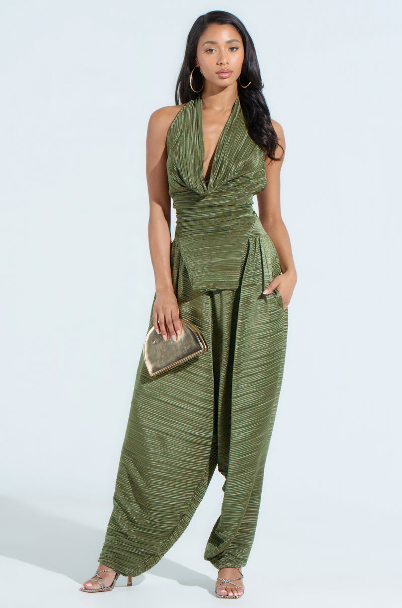 A woman confidently wears the CROIX DROP CROTCH PLISSE PANT, an olive-green halter jumpsuit with stylish drop crotch pants. Holding a gold clutch, she's adorned with large hoop earrings and strappy heels, her long hair cascading over one shoulder.