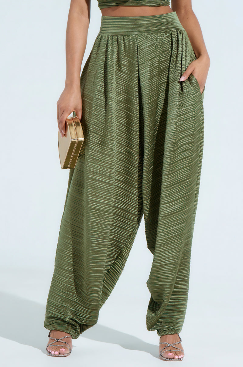 A person wearing the CROIX DROP CROTCH PLISSE PANT in olive green holds a gold clutch. The high-waisted, drop-crotch pants are paired with strappy heels against a plain, light gray background.
