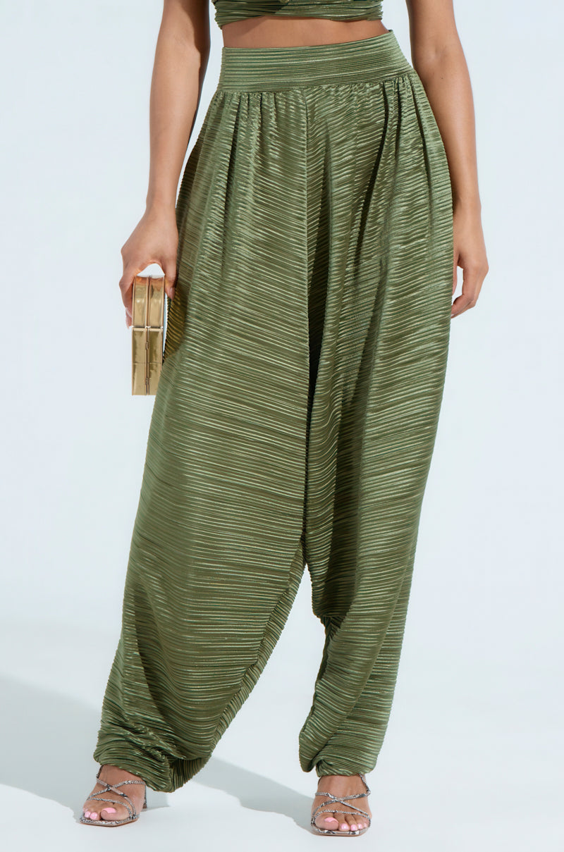 A person wearing CROIX Drop Crotch Plisse Pants in textured green and transparent heels holds a beige clutch, photographed from the waist down against a plain background.