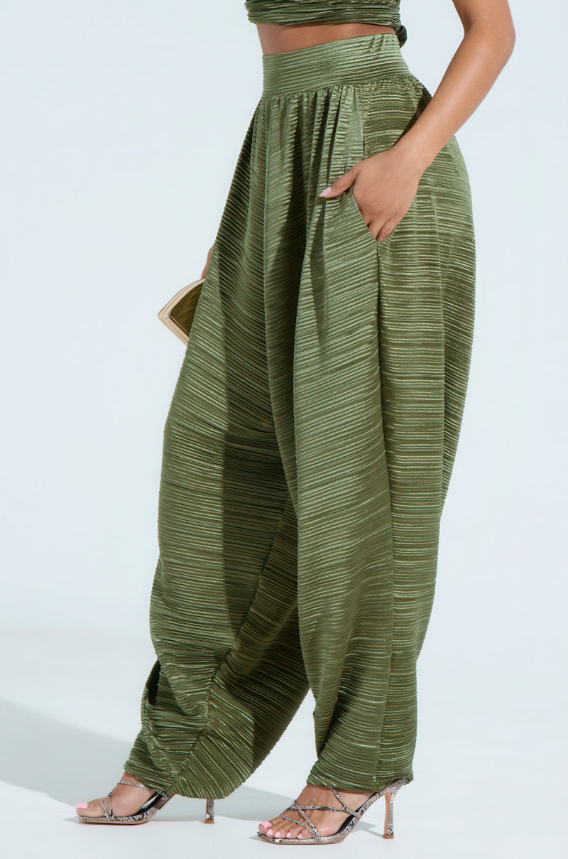 The person wears green CROIX DROP CROTCH PLISSE PANT, featuring high-waist and wide-leg with horizontal textured lines. One hand in a pocket, they hold a small clutch and pair the look with strappy high-heeled sandals against a plain white background.