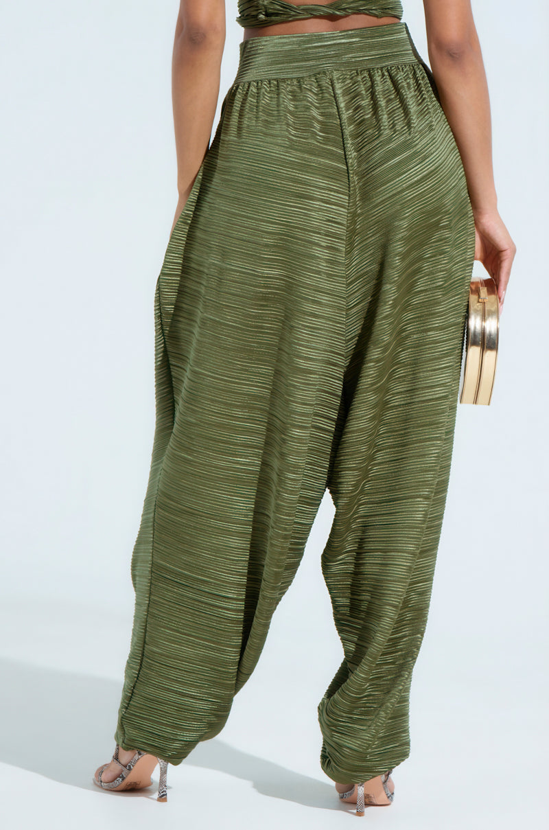 A person stands on a light background, wearing loose-fitting CROIX DROP CROTCH PLISSE PANT in textured green. They hold a gold clutch and wear strappy high heels.