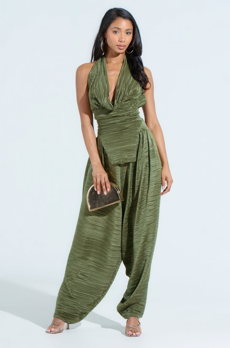 A woman models the CROIX DROP CROTCH PLISSE PANT, an olive-green jumpsuit with a draped fabric and halter neck. She accessorizes with hoop earrings, strappy sandals, and a metallic clutch. The light background enhances the stylish ensemble and highlights its unique silhouette.