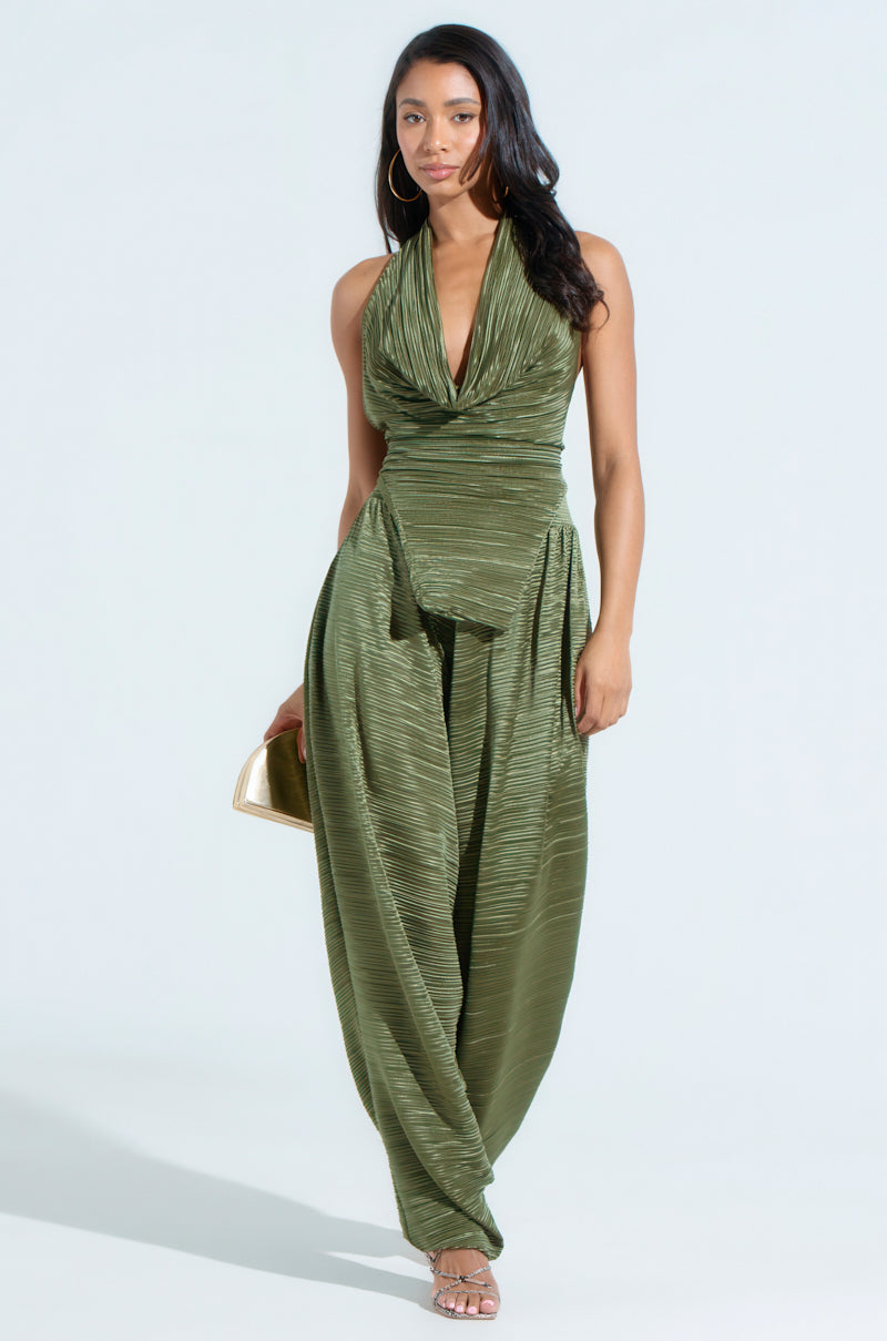 Against a plain backdrop, a woman wears a sleeveless green jumpsuit with a draped neckline and wide legs similar to the CROIX DROP CROTCH PLISSE PANT. She holds a gold clutch, wears hoop earrings and sandals, and her long dark hair falls in loose waves.