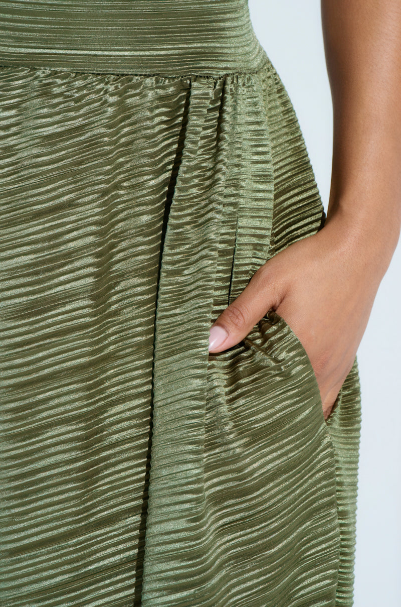 In a close-up, the right hand of a person delicately holds the fabric of the CROIX DROP CROTCH PLISSE PANT, highlighting its pleated, olive-green texture. The neatly manicured nails elevate the scene's elegance.