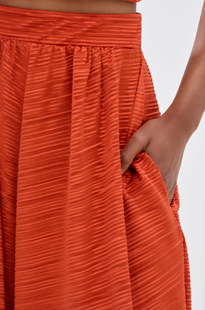 A close-up shows a person wearing shiny, ribbed MAYAH WIDE LEG PLISSE PANT IN RED, pleated with a hand casually resting in the pocket. 5