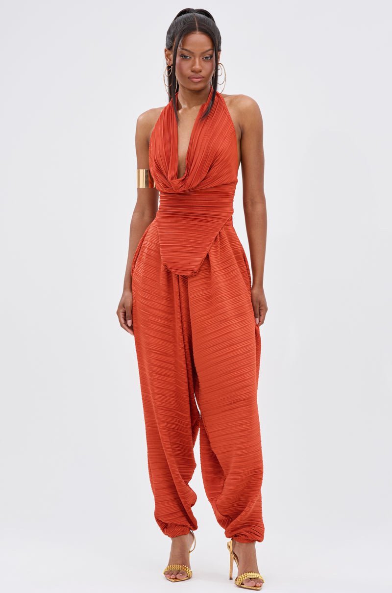 A woman stands against a white background wearing orange pleated halter-neck jumpsuit with a draped cowl neckline, wide gathered pants inspired by the MAYAH COWL NECK PLISS SLEEVELESS TOP IN RED, gold heels, a cuff bracelet, and long earrings. 5