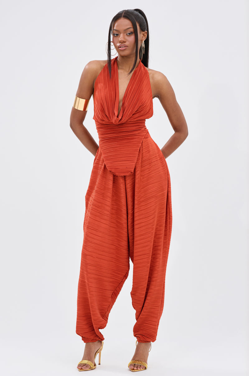 A woman stands confidently in the MAYAH COWL NECK PLISS SLEEVELESS TOP IN RED, featuring a draped cowl neckline. She completes her look with gold high heels, large hoop earrings, and a bold gold cuff bracelet. 6