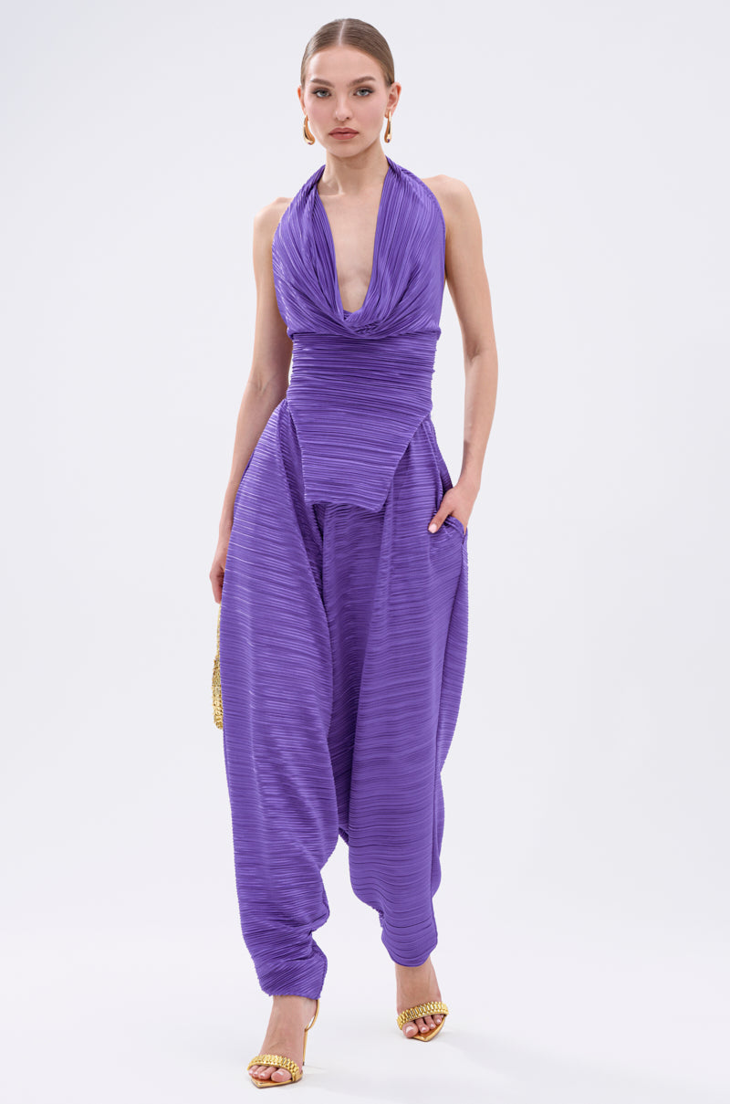 A woman stands against a plain white background wearing the MAYAH Wide Leg Plisse Pant in purple, styled with gold high-heeled sandals and bold earrings, paired with a vibrant halter-neck top featuring a draped, plunging neckline. 0