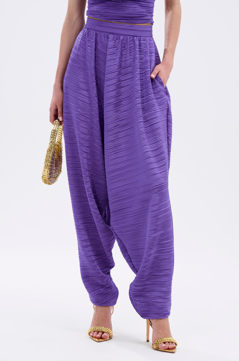 A person stands against a plain white background wearing the MAYAH WIDE LEG PLISSE PANT IN PURPLE, holding a gold beaded clutch and wearing gold high-heeled sandals. 1