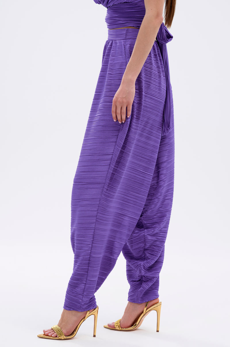 A woman wears the MAYAH WIDE LEG PLISSE PANT IN PURPLE with gold high-heeled sandals and a hint of a matching purple top, set against a plain white background. 3