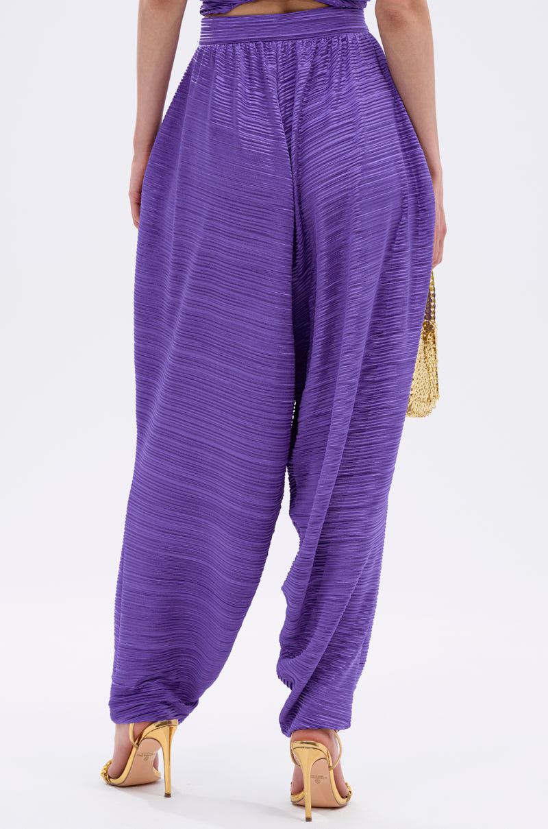 A person is seen from the back wearing the MAYAH WIDE LEG PLISSE PANT IN PURPLE with gold high-heeled sandals and a small gold bag, standing against a plain white background. 4