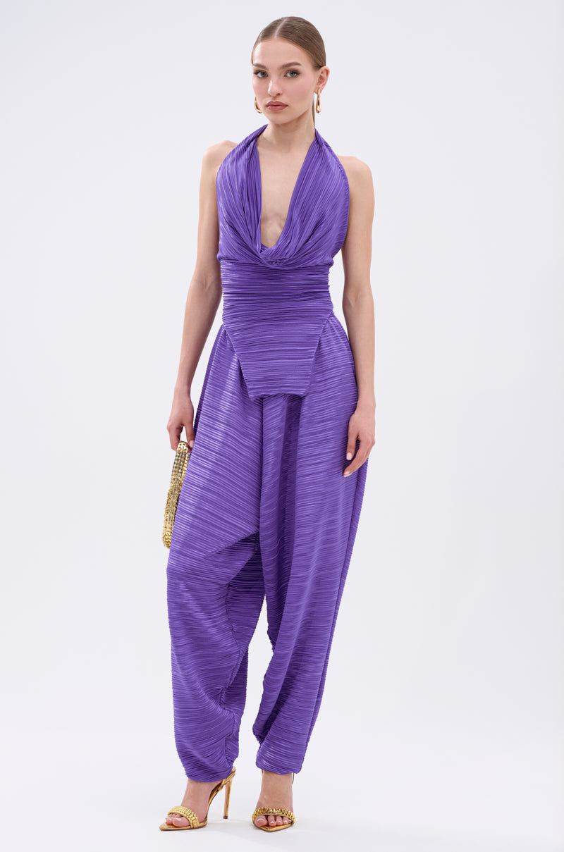 A woman wears the MAYAH Wide Leg Plisse Pant in purple, styled with a gold clutch and gold high-heeled sandals, standing against a plain white background. 6