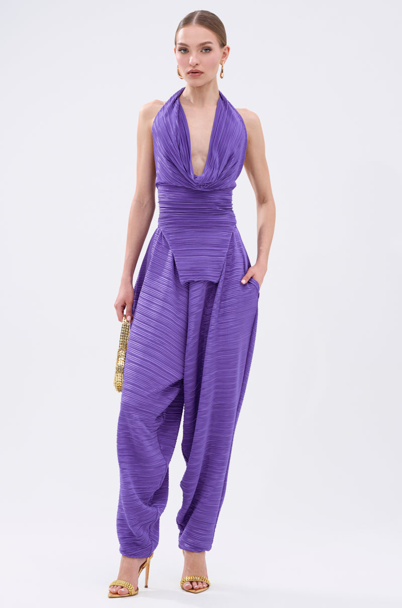 A woman poses against a white background wearing the MAYAH COWL NECK PLISSE SLEEVELESS TOP IN PURPLE, paired with wide plissé pants, gold high-heeled sandals, hoop earrings, and holding a gold beaded clutch. 6