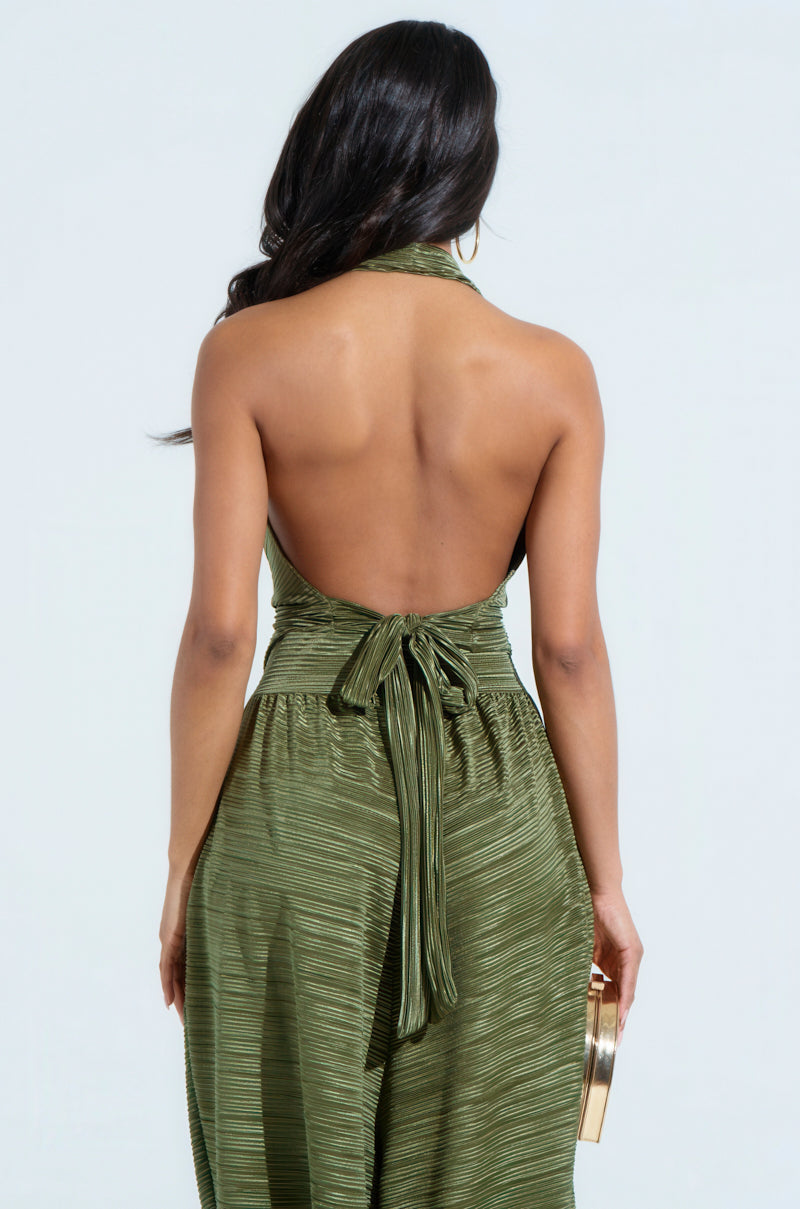 Wearing a CROIX COWL NECK PLISSE HALTER TOP in green, the woman, with long dark hair elegantly falling over her shoulders, holds a gold clutch. The top features an open back design and tied waist, set against a plain light gray backdrop.