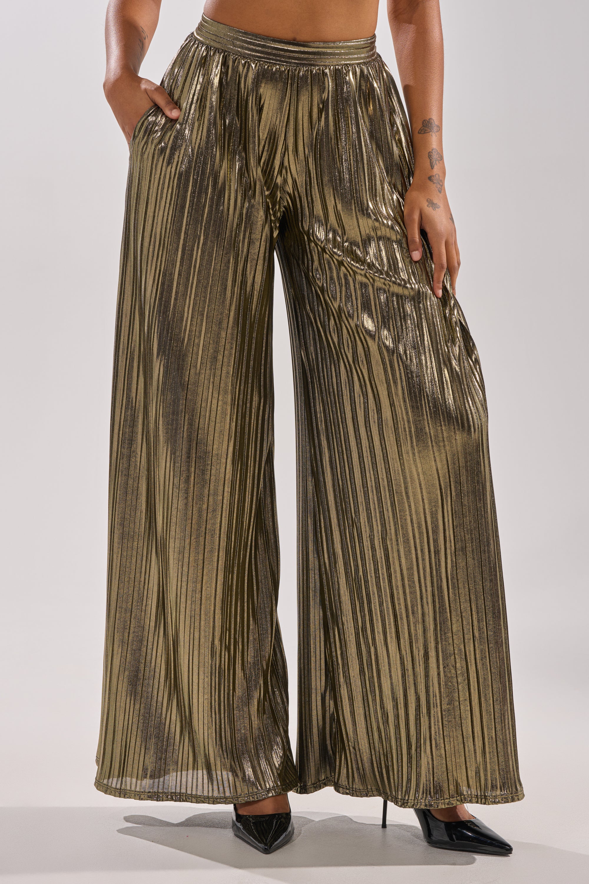A person models the BEST DRESSED PALAZZO PANT—high-waisted, metallic pleated wide-leg pants—with hands in pockets, paired with black pointed-toe heels. Only the lower body is visible. 0