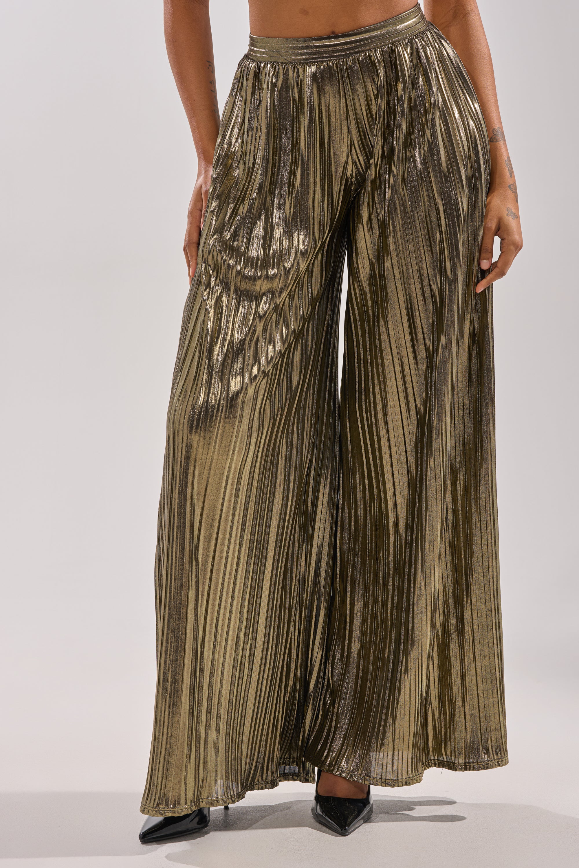 A person wearing the BEST DRESSED PALAZZO PANT—shiny, high-waisted, metallic, pleated wide-leg pants—paired with black pointed-toe heels stands against a plain light background. Only the lower half is visible. 1
