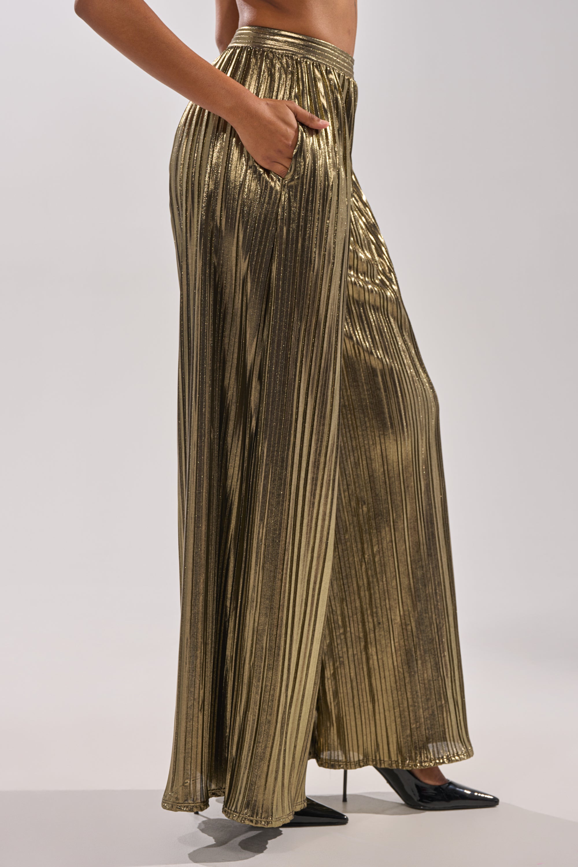 A person wearing the BEST DRESSED PALAZZO PANT—metallic, pleated, wide-leg style with a hand in the pocket—paired with pointed black heels, photographed from the side against a plain background. 2