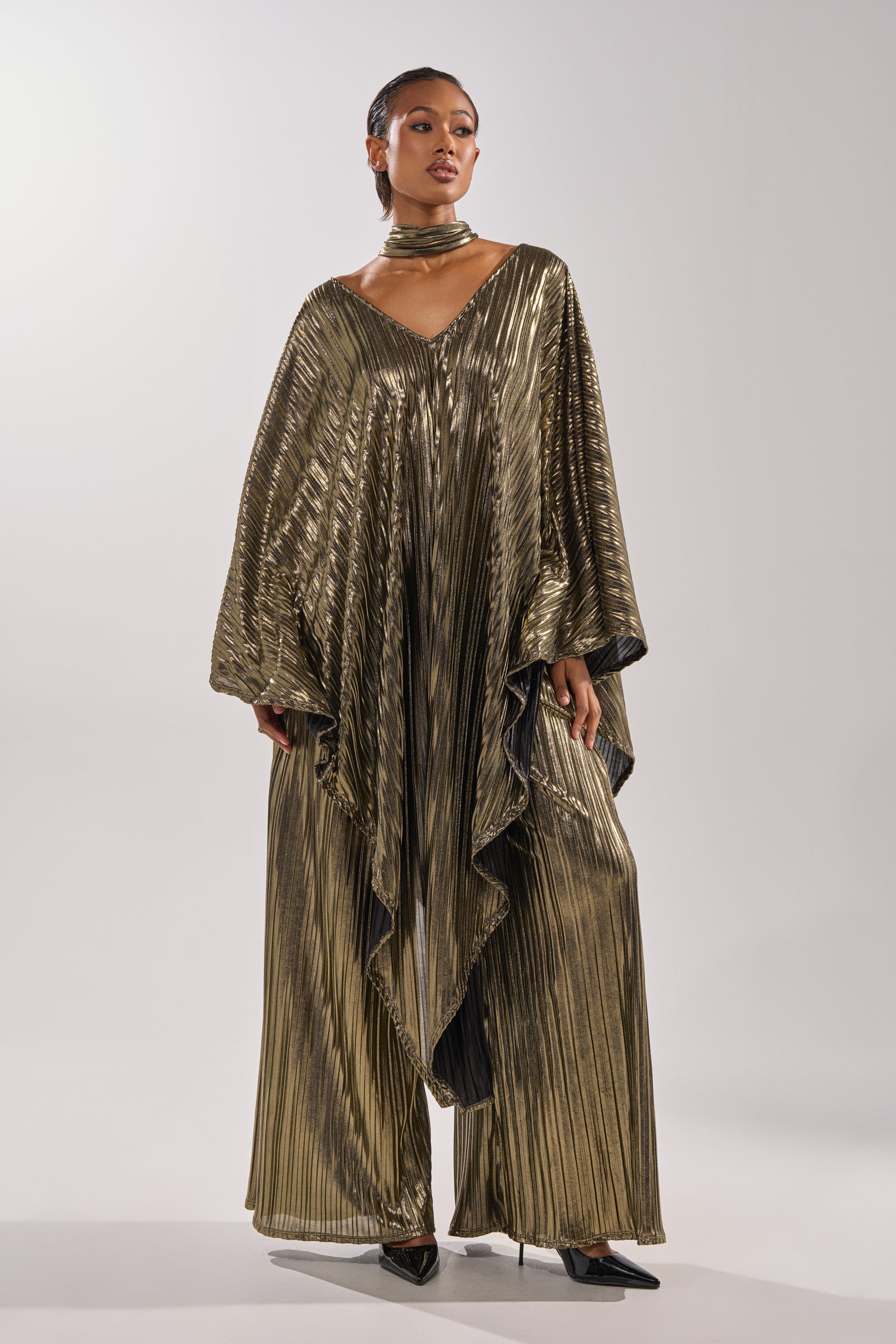 A woman wears the BEST DRESSED BLOUSE—a flowing, metallic pleated top with wide sleeves—paired with pants, a matching choker, and black pointed heels, posing stylishly against a plain light background. 0